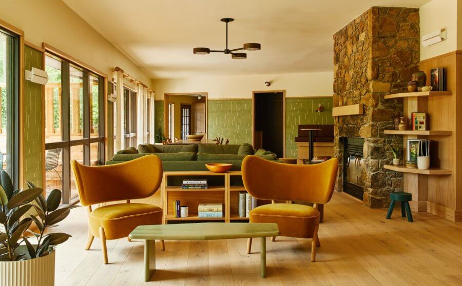 23 New Boutique Hotels in the South - 5 In a style reminiscent of new boutique hotels, this modern living room features mustard armchairs, a green sofa, and a wooden coffee table beside a stone fireplace. Large windows line one side while lush indoor plants add vibrant greenery to the inviting space.