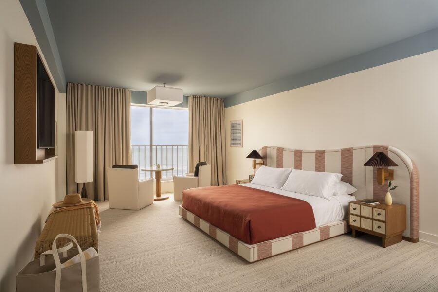 23 New Boutique Hotels in the South - 13 This stylish room in a new boutique hotel features a striped bed with a red blanket, flanked by two nightstands and a flat-screen TV. A window with curtains opens to reveal a balcony view, while a small table and two chairs invite relaxation.