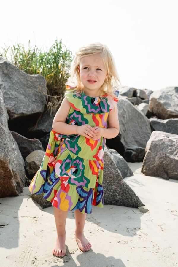 A little blond girl stands on the beach in a dress.