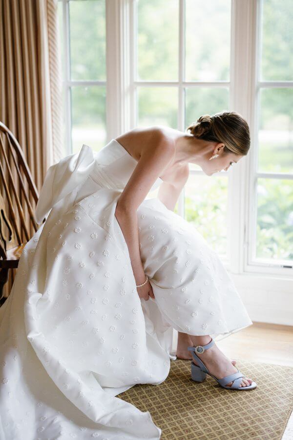 A North Carolina Wedding Dressed in Blues, Bows, and Bojangles - 9 A bride is putting on her shoes in front of a window at a Linville Wedding.