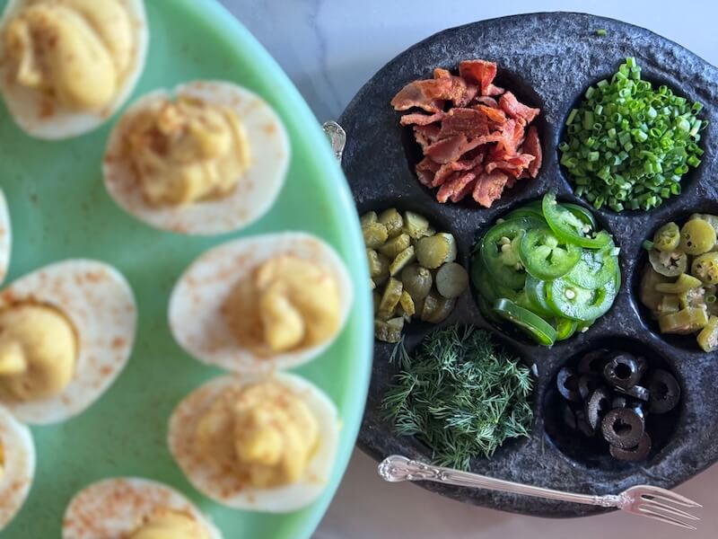 A deviled egg bar featuring a platter of deviled eggs accompanied by various garnishes including chives, pickles, and herbs.