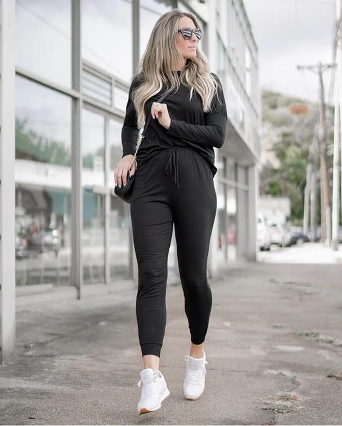 A woman in a black outfit with white sneakers is standing on a sidewalk.