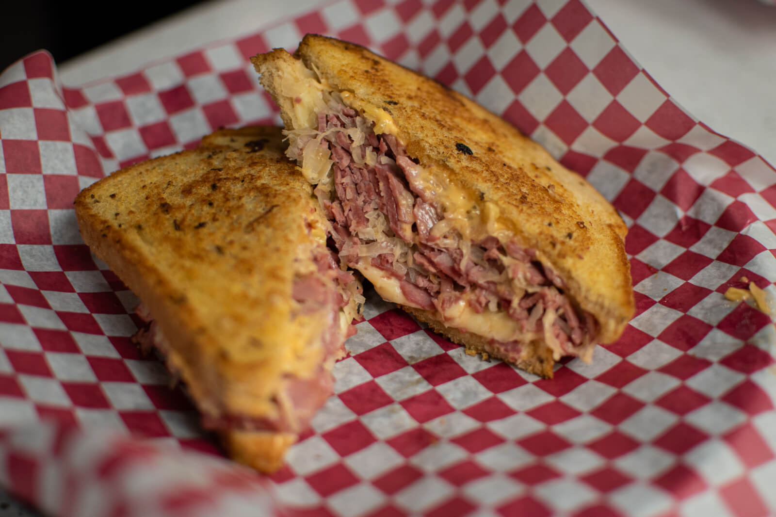 A reuben sandwich with melted cheese and corned beef on toasted bread, served on a red and white checkered paper.