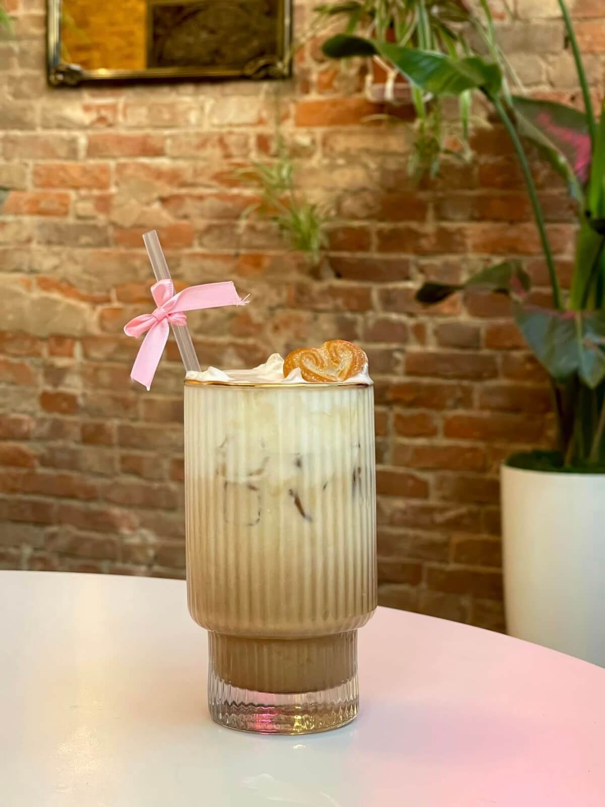 Iced coffee garnished with whipped cream and a cookie, served with a straw tied with a pink ribbon, against a brick wall backdrop.