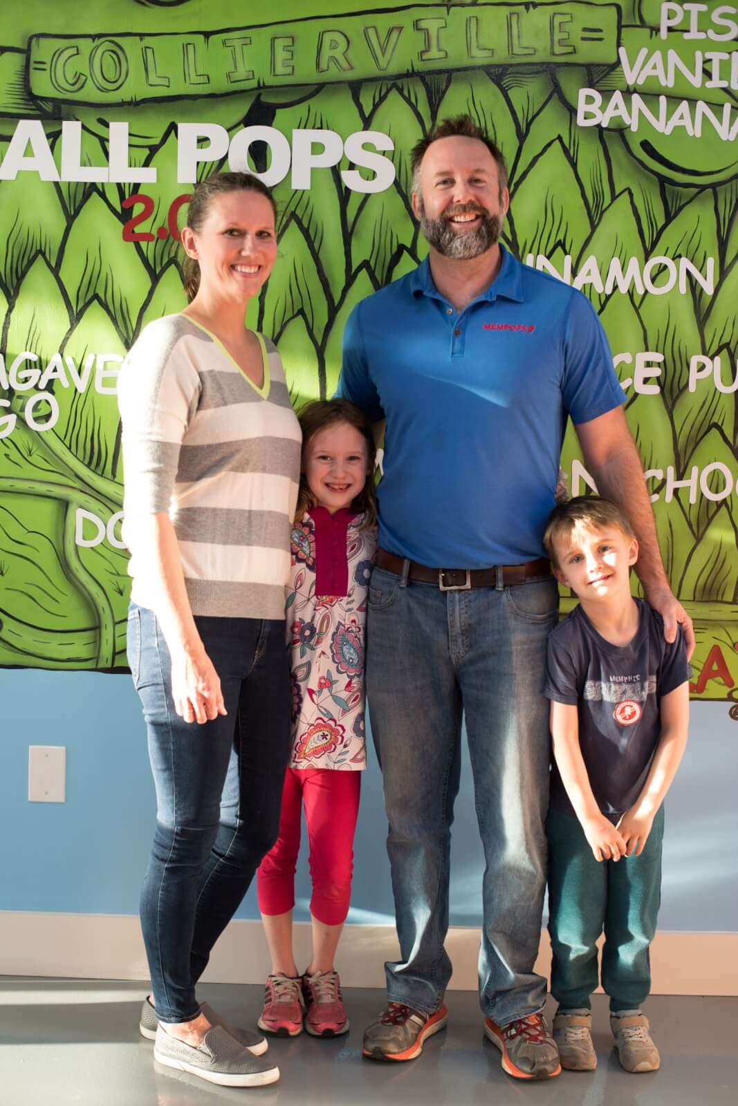 A family of four smiling in front of a colorful wall with the words "all pops" displayed.