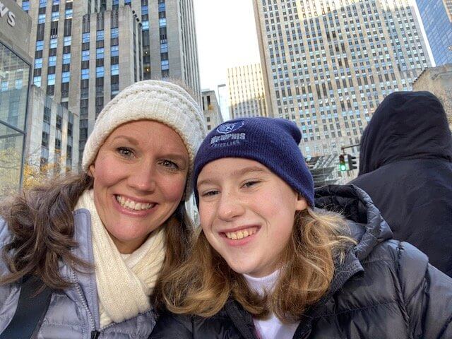 Two people smiling for a selfie outdoors, with city buildings in the background.
