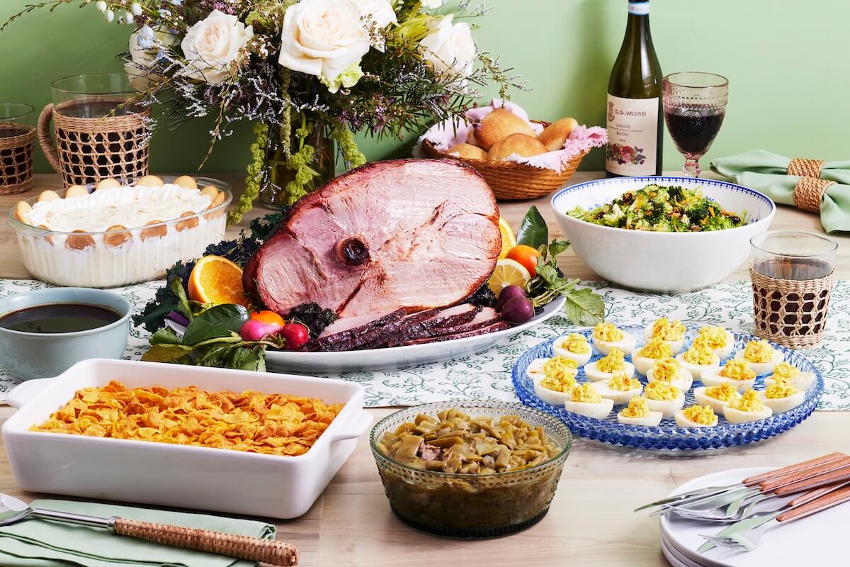 A table set for a Nashville Easter brunch features a sliced ham centerpiece, surrounded by deviled eggs, casserole, green beans, salad, bread rolls, wine, and dessert. White flowers add a touch of elegance to the background.