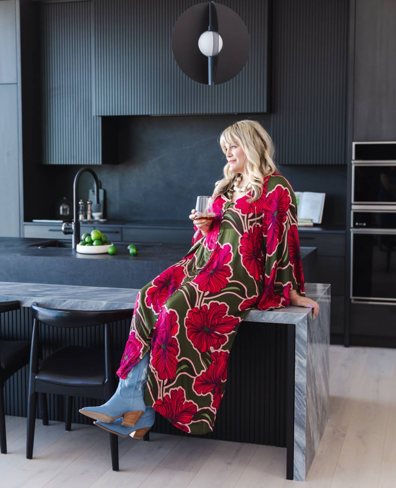 A woman in a springtime floral dress sits on a kitchen counter, holding a drink.