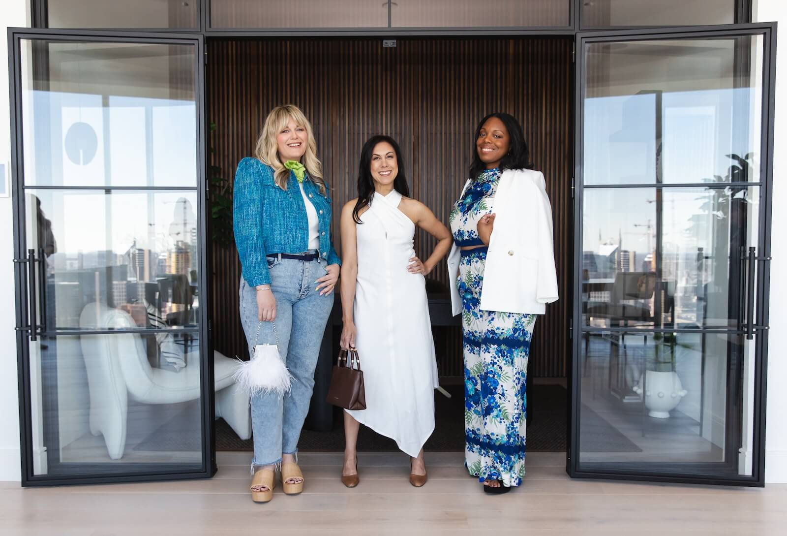 Three women posing together, dressed in professional spring attire, inside a modern office building.