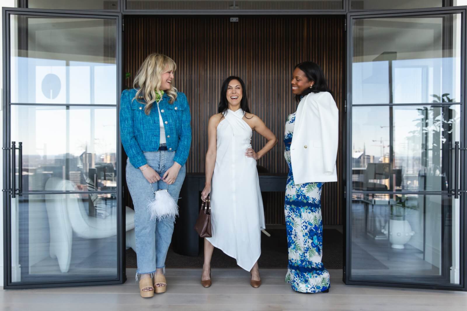 Three women, dressed in what to wear for spring, laughing and conversing in a modern office setting.