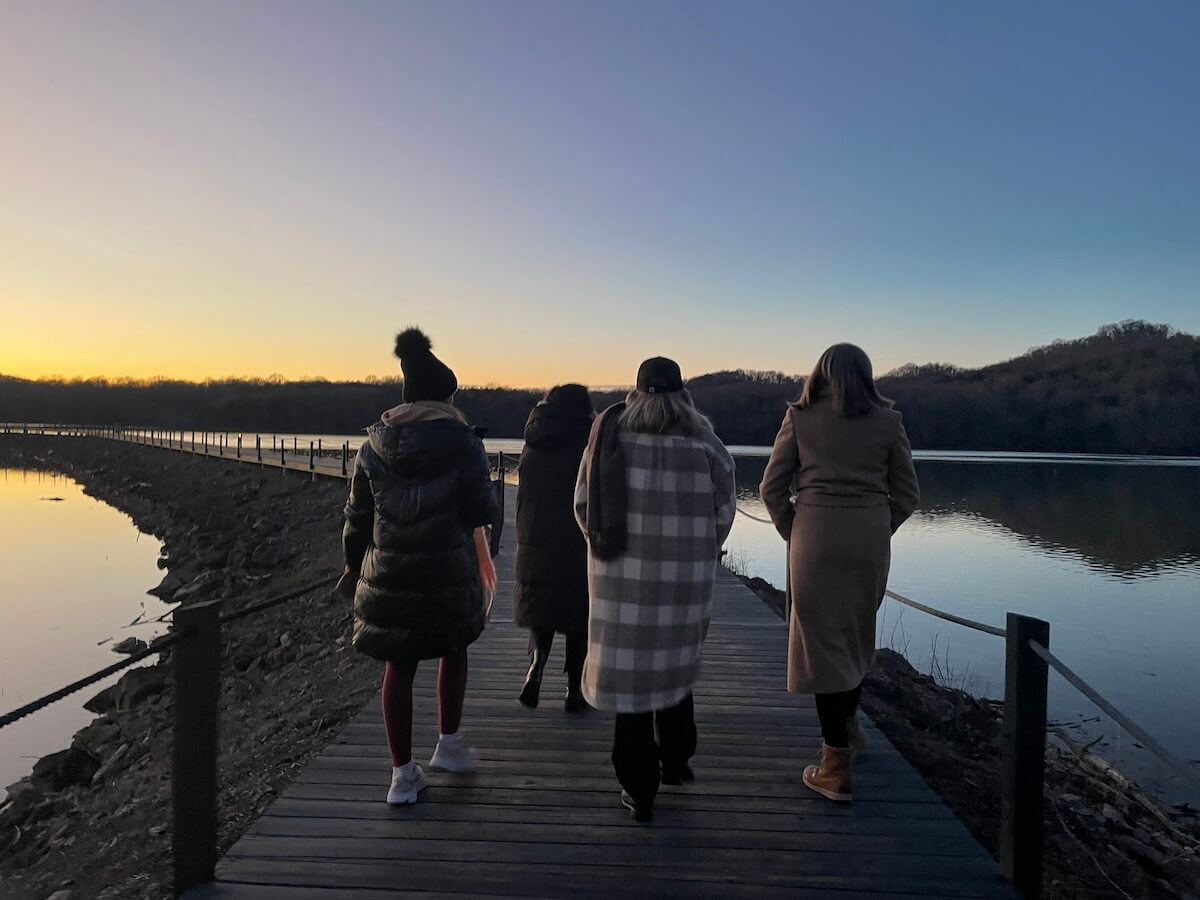 Group of people walking on a wooden dock at dusk with a calm lake on one side and a treelined horizon in the background.