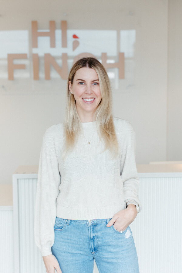 A woman wearing jeans and a sweater standing in front of a hi finch sign.
