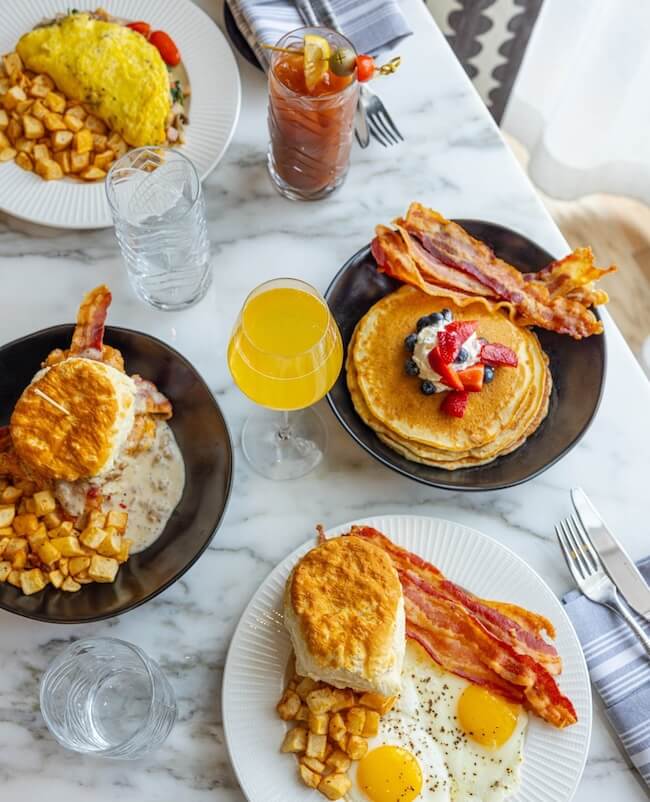 A brunch in Memphis offers a spread of delectable breakfast dishes such as pancakes with fruit, bacon, eggs, biscuits with gravy, home fries, alongside a refreshing Bloody Mary and a lively mimosa.