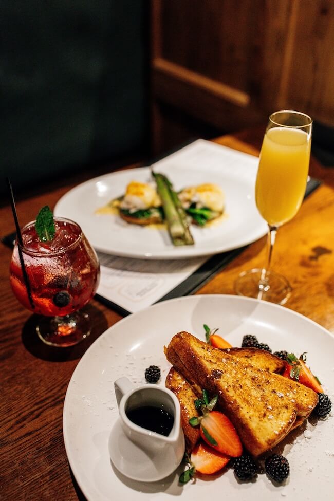 French toast with strawberries and blackberries, a small jug of syrup, a glass of orange juice, and a berry-garnished cocktail grace the wooden table during brunch in Memphis. A blurred dish peeks from the background.