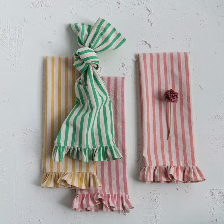 Striped fabric napkins with ruffled edges in vibrant yellow, green, and pink are artfully arranged on a textured white surface. A dried flower graces one of the pink napkins, offering a charming touch to this delightful March find.