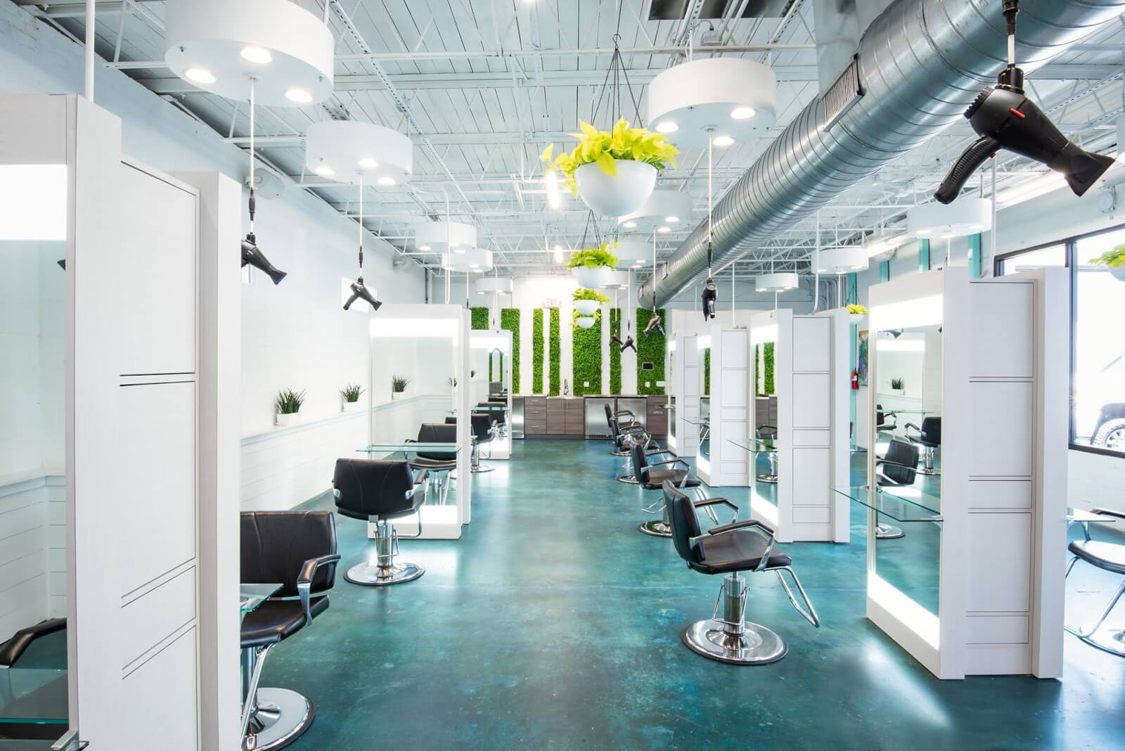 A modern hair salon in Birmingham, featuring styling stations, black chairs, large mirrors, overhead hair dryers, and green plant decor. Bright lighting and blue flooring create an open and airy atmosphere at one of the best spots for top-notch spa services in town.