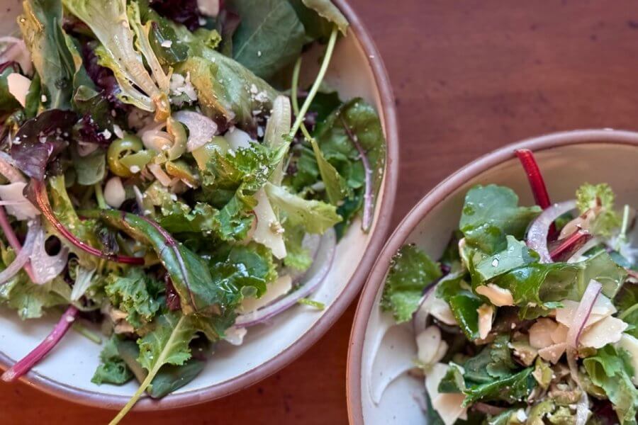 Two bowls of fresh mixed green olive salads with assorted leaves, herbs, and grated cheese on a wooden table.