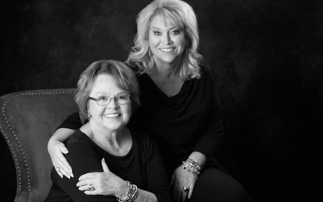 Carolyn Kelley sits on a chair with daughter Cathy Kelley sitting behind her, both in black tops against a dark background.