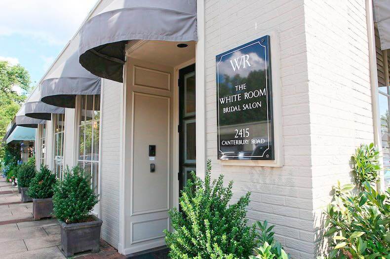 Exterior view of The White Room bridal salon with a sign, entrance door, and plants along the sidewalk.