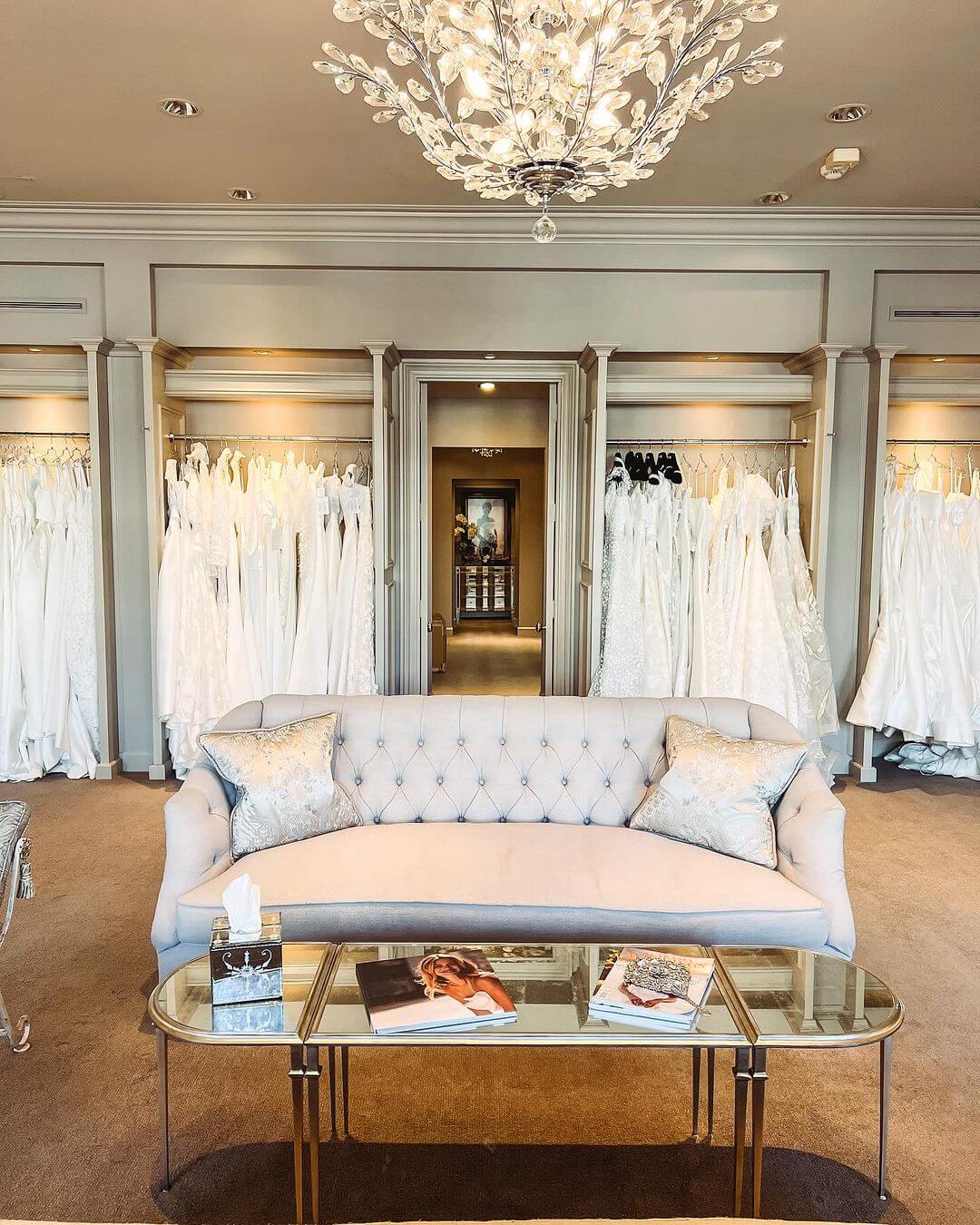 Elegant bridal shop interior featuring a plush sofa, glass coffee table with magazines, and rows of white wedding dresses at The White Room.