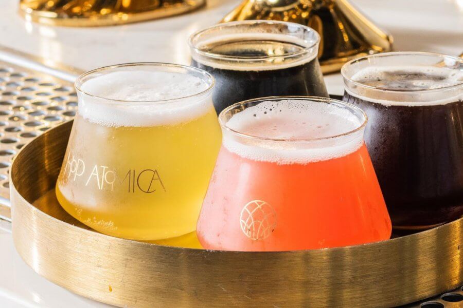 A flight of various beers in glasses on a golden tray, each with a unique color from pale yellow to dark brown