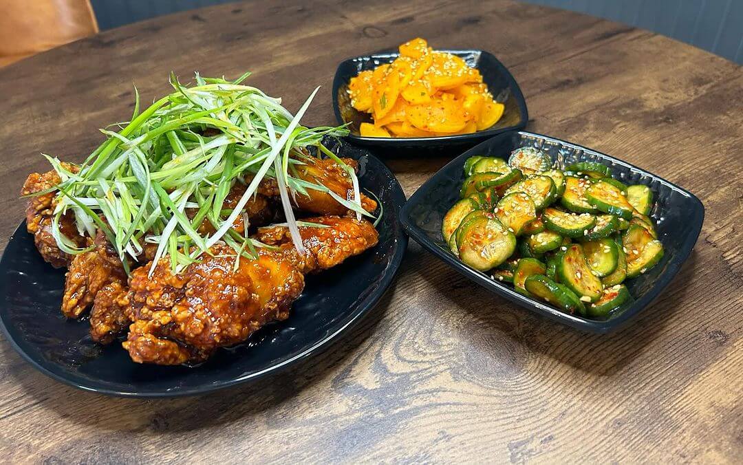 A wooden table with three dishes: glazed chicken topped with scallions, a bowl of spicy sliced cucumbers, and a bowl of marinated orange segments.