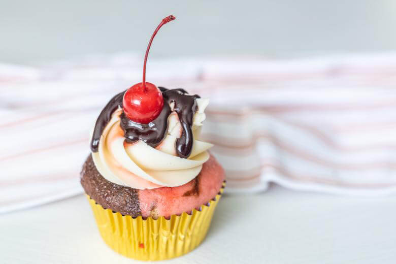 A colorful cupcake with pink and chocolate layers, topped with white frosting, chocolate syrup, and a cherry on a striped surface.