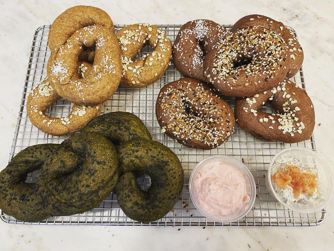 Various bagels with different toppings, including plain, sesame, and spinach, displayed on a cooling rack with two types of cream cheese on the side.