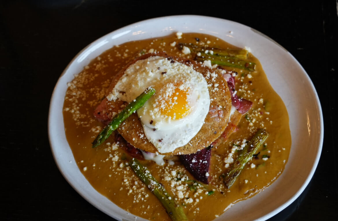 A plate of food featuring stacked pancakes topped with a fried egg, bacon, asparagus, and a drizzle of syrup.