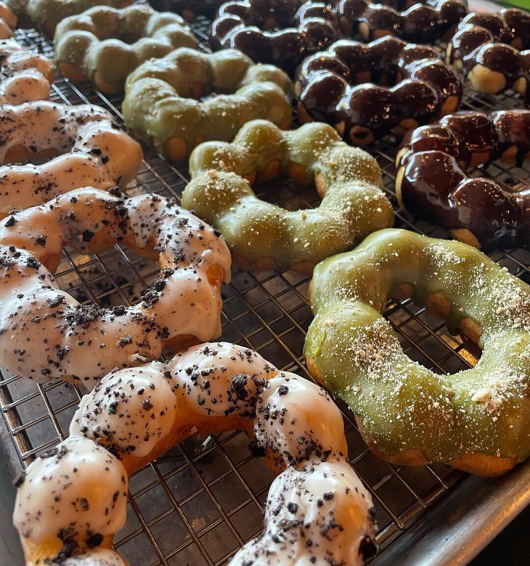 Assorted doughnuts on a cooling rack, including white with black specks, and green and chocolate glazed varieties.
