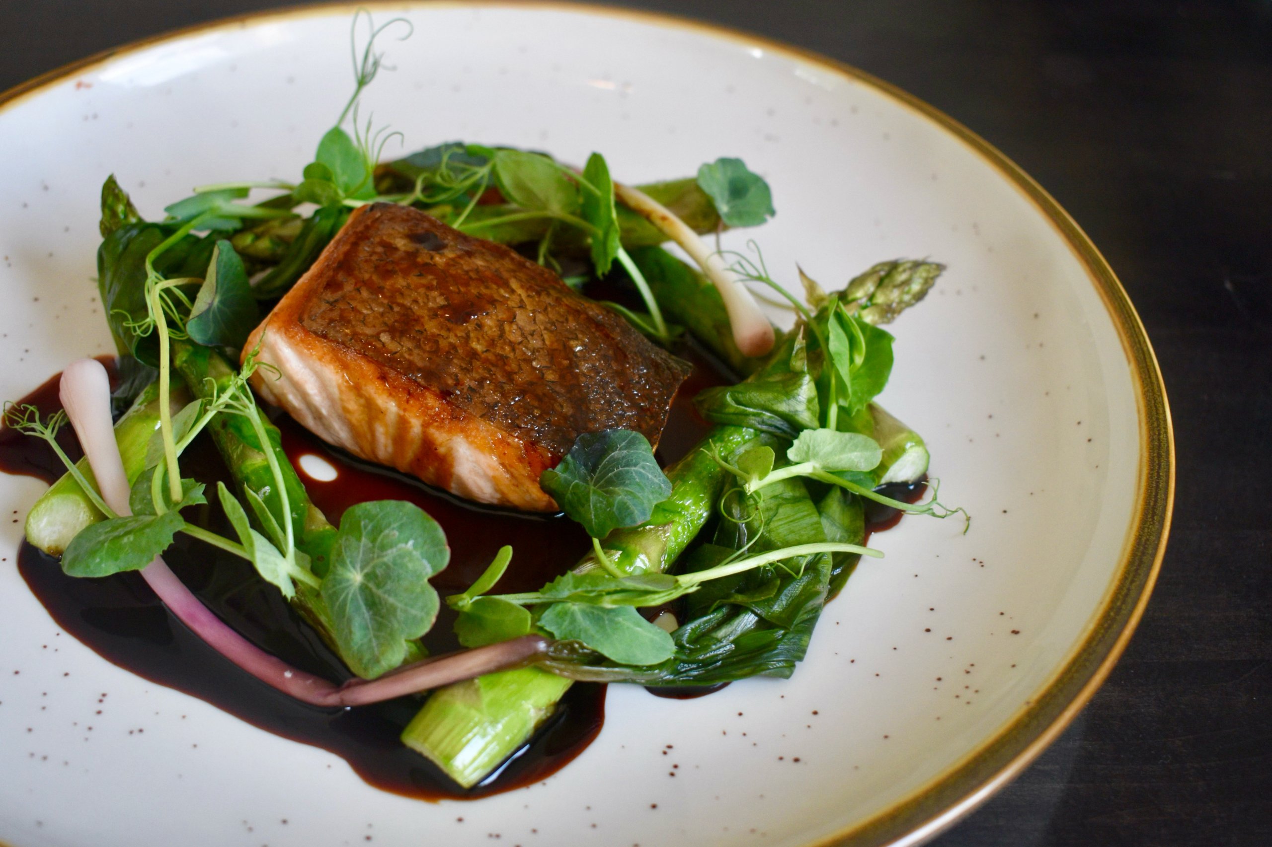 Pan-seared salmon fillet served on a bed of green vegetables and garnished with herbs, presented on a white plate with gold rim.
