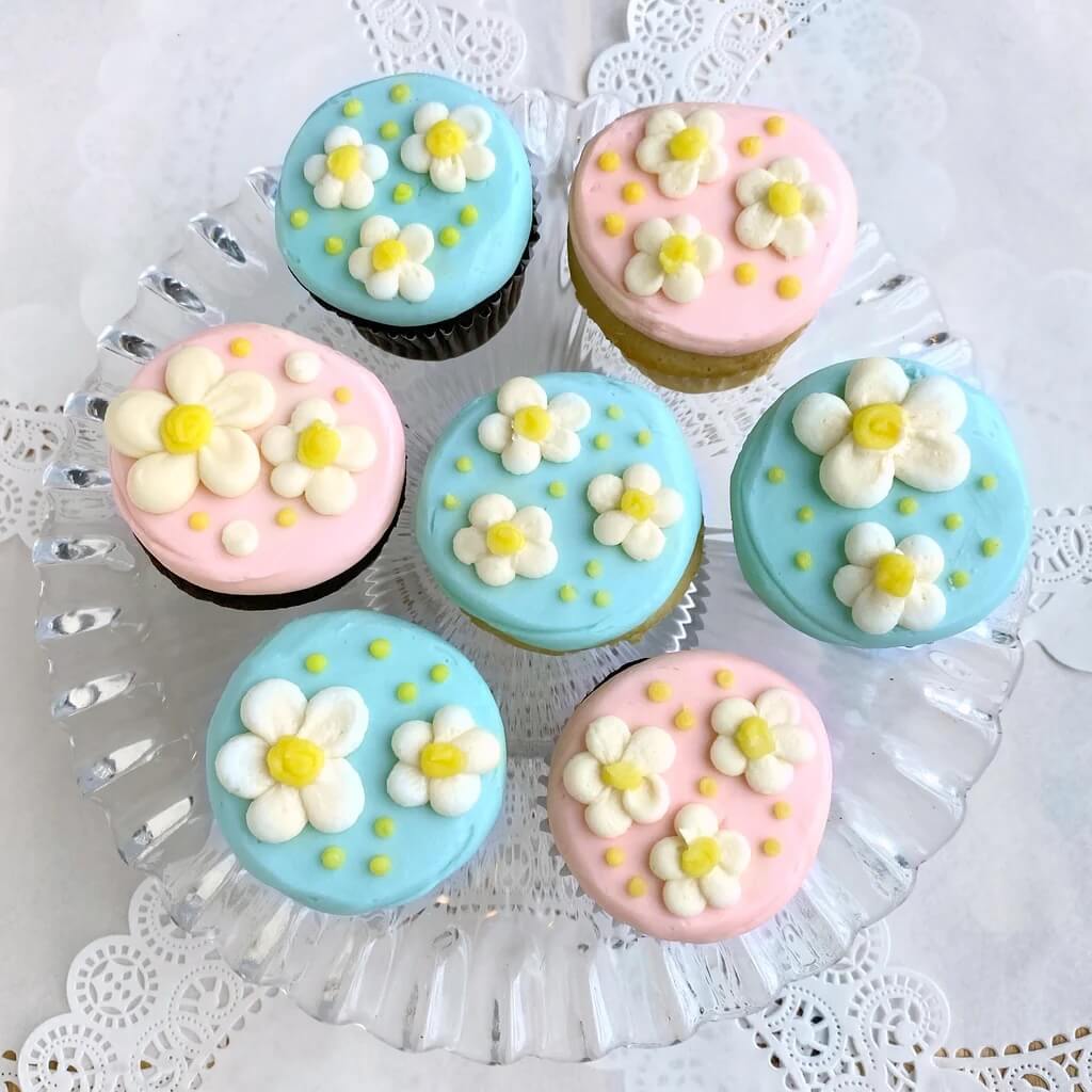 Eight cupcakes with pastel pink and blue frosting, decorated with white and yellow fondant flowers, displayed on a lace doily.