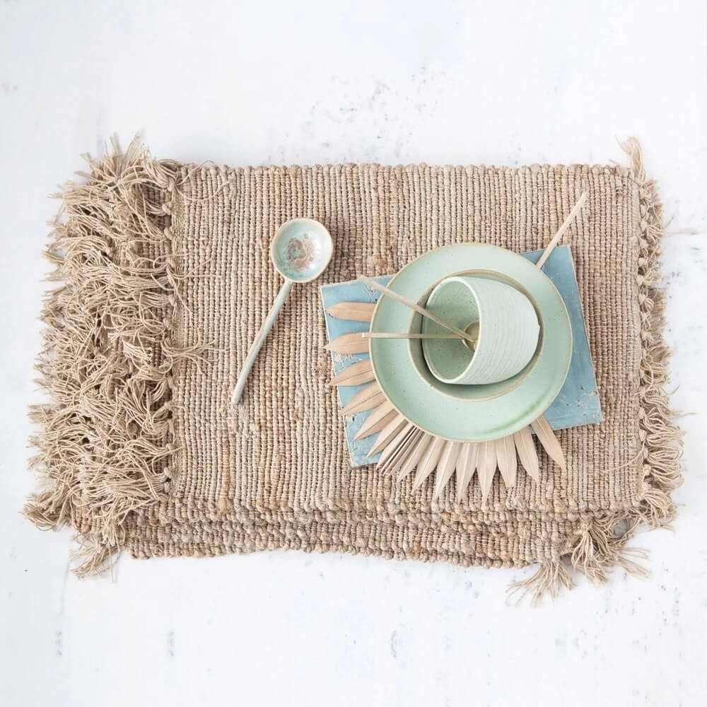Top view of a neatly set dining place on a textured mat featuring a ceramic plate, bowl, cup, and silverware, all arranged on a natural, fringed table runner.