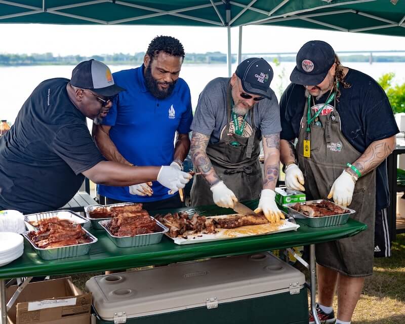 Four men collaborating to prepare barbecue outdoors, standing around a green table with plates of meat and barbecue tools.