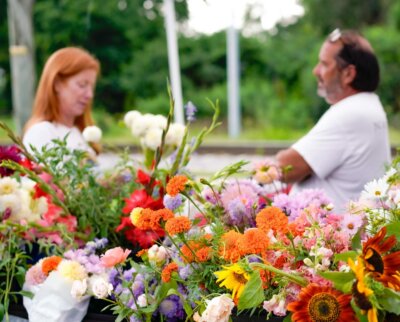 Two people stand behind tables displaying a variety of colorful cut flowers outdoors at one of the lively Nashville farmers markets, with greenery in the background.