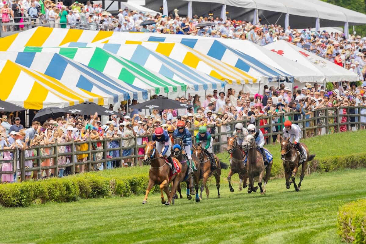 Several jockeys ride racehorses on a grassy track as a large crowd watches from behind a wooden fence and colorful striped tents—an exciting scene often found at Nashville events in May.
