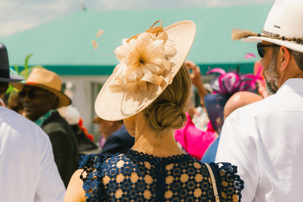 A woman wearing a decorative beige hat and navy lace dress stands among a crowd at an outdoor Southern event in May; people around her also wear hats.