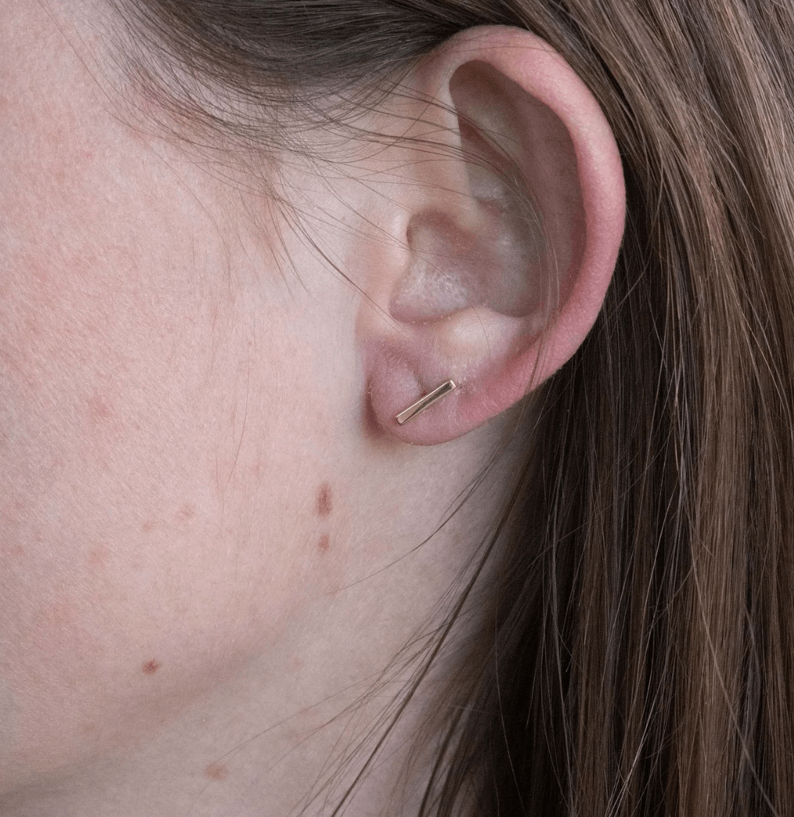 Close-up of a woman's ear wearing a small gold bar earring, with visible brown freckles on her cheek.