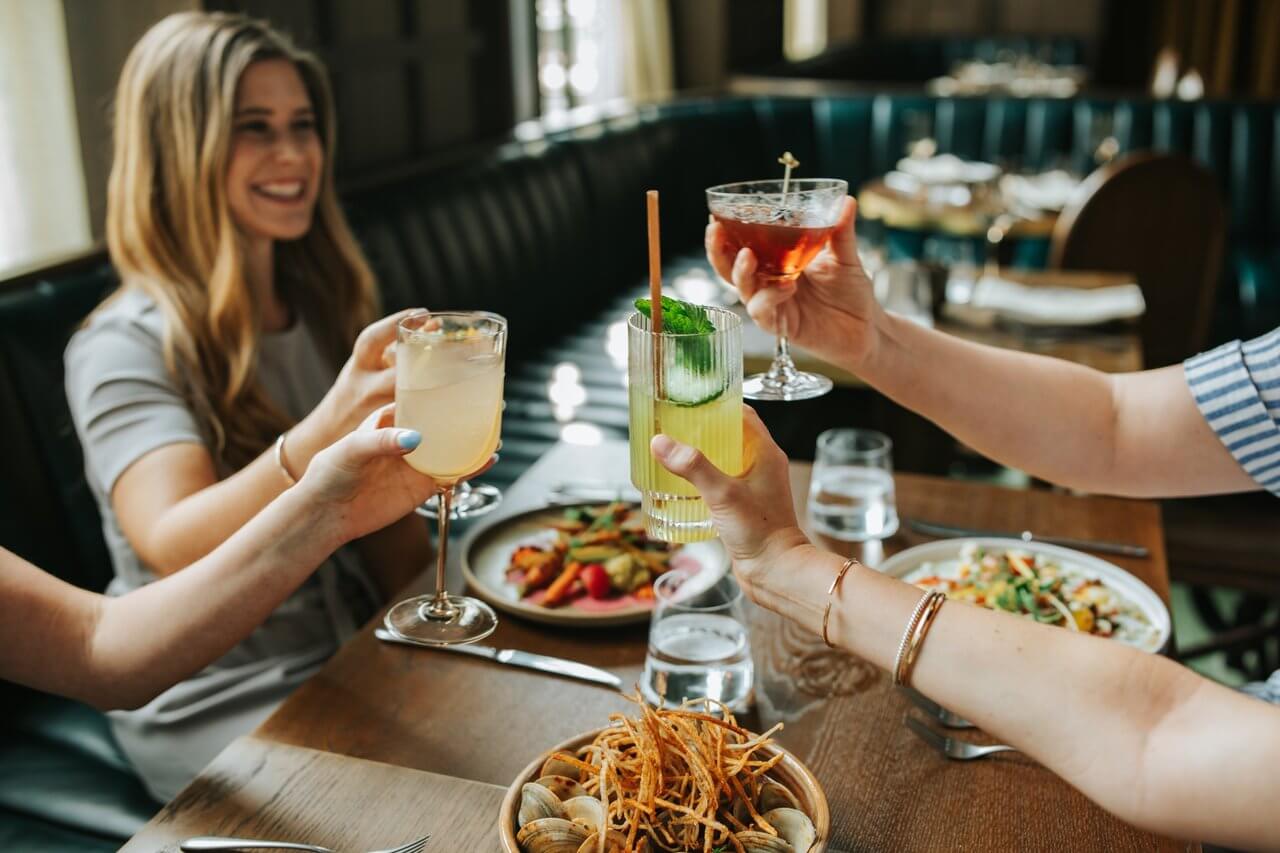 Group of people toasting with cocktails over a restaurant table with food.