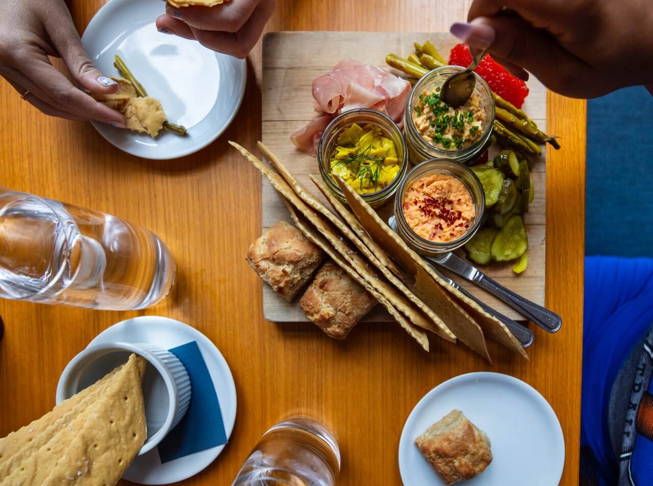 Overhead view of a charcuterie board with assorted meats, pickles, dips, and breadsticks, with people reaching in to share the meal.