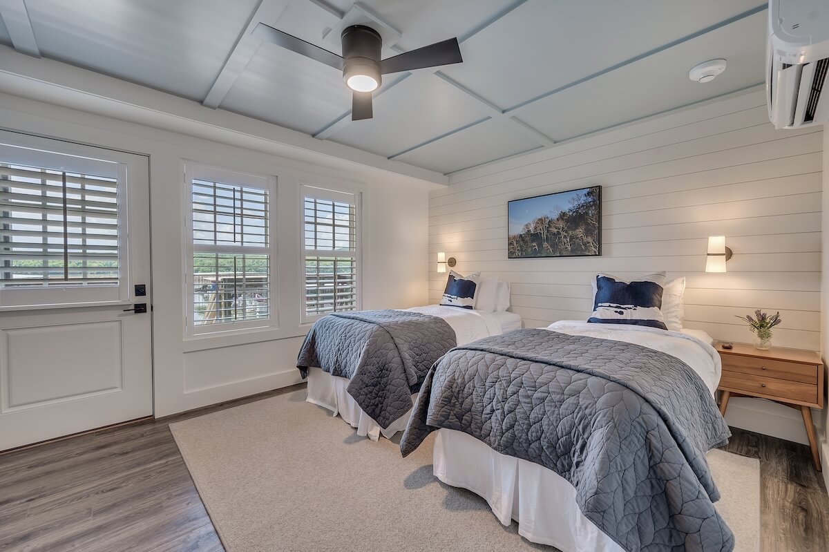 A bright twin bedroom in Wildwood Resort with a coastal theme, featuring two beds with navy and white bedding, a ceiling fan, and shuttered windows.