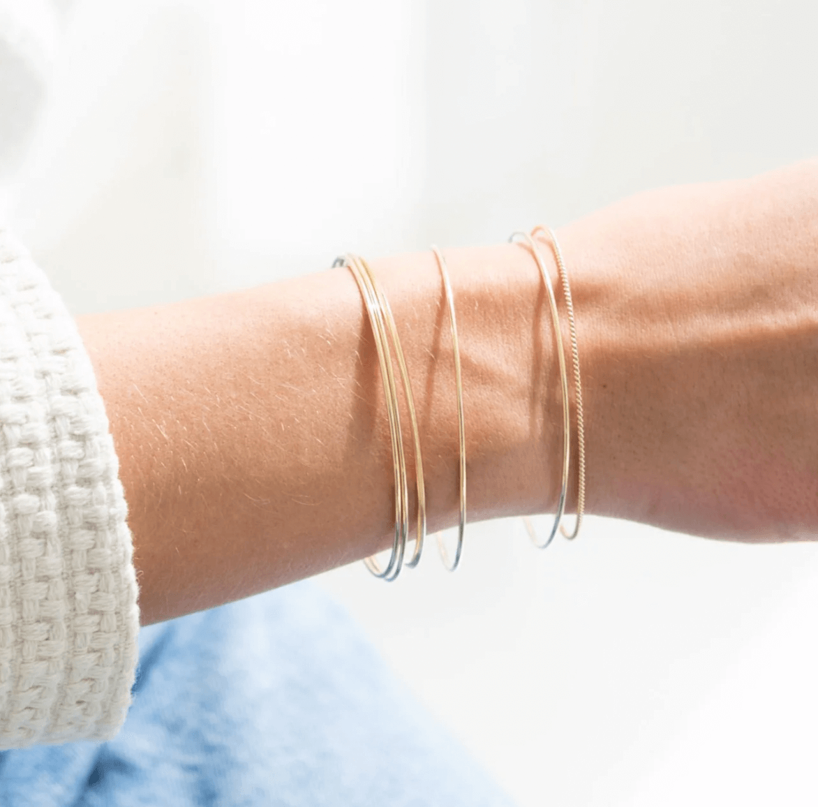A person's wrist adorned with four thin, gold bangle bracelets, shown against a soft white background.
