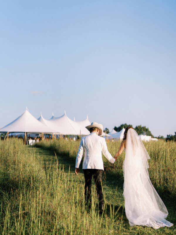 A couple holding hands and walking towards a wedding tent in a field.