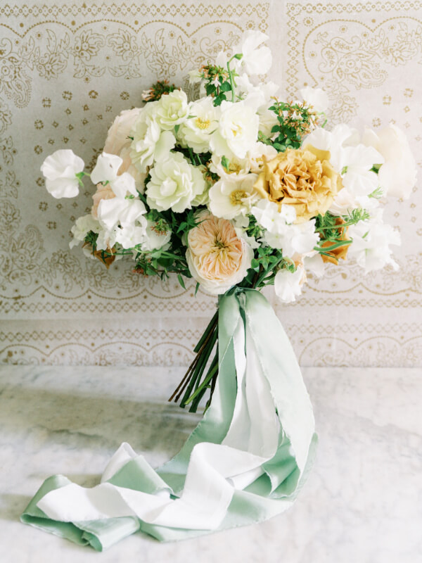 An elegant bouquet of white and pale yellow flowers tied with a soft mint green ribbon against a patterned backdrop.