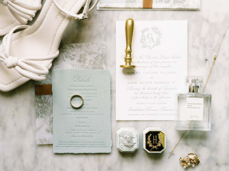 Wedding invitation suite with elegant calligraphy, a pair of white bridal shoes, perfume bottle, and accessorizing jewelry on a marble surface.