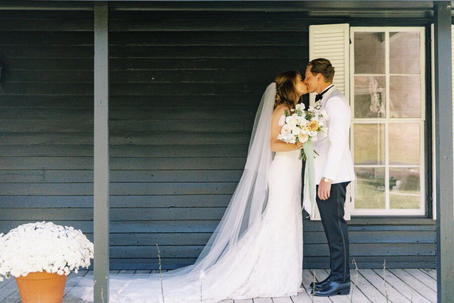A bride and groom kissing in front of a dark wooden wall with a window and white shutters.