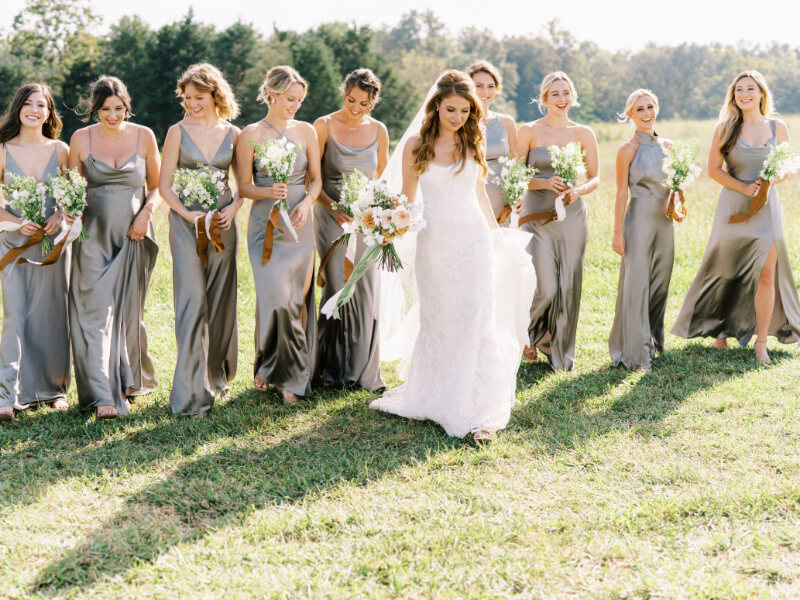 A bride walks confidently ahead of her bridesmaids who are dressed in coordinated gowns, holding bouquets in an outdoor setting.