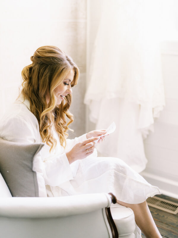 A woman in a white robe sitting by a window, smiling as she reads a card.