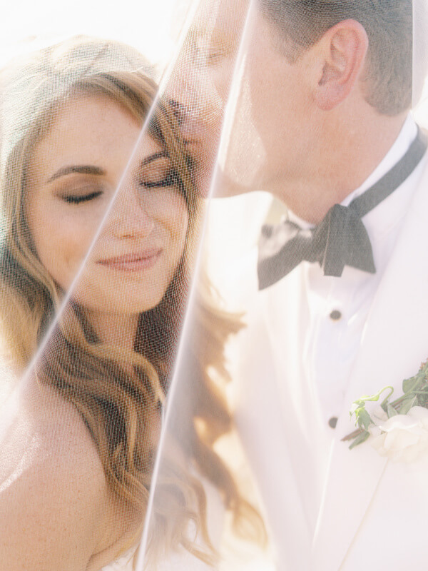 A groom kissing his bride on the forehead during a sunlit wedding ceremony.