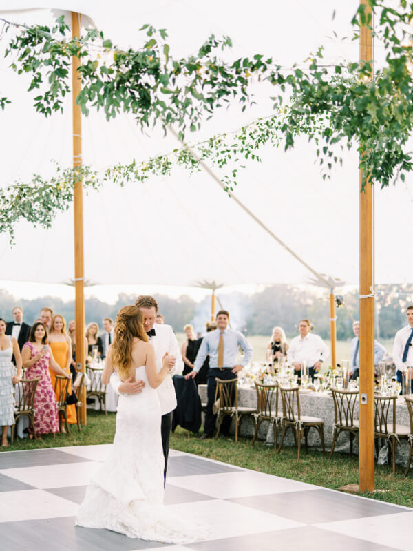 A bride and groom share their first dance outdoors with guests looking on.